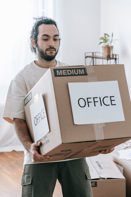 A man with dark, curly hair and a beard is indoors, holding a medium-sized cardboard box labeled 'OFFICE' and 'MEDIUM' in black print, during a home or office relocation process. The box is sealed wit