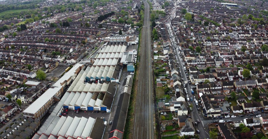 Aerial view of a residential area in Chessington, showing a railway line running vertically through the centre of the image alongside a large industrial warehouse complex with multiple sloped, semi-transparent roof sections. Surrounding the warehouse are numerous terraced and semi-detached houses with small gardens, driveways, and tightly packed streets, all under overcast sky lighting. Visible cars are parked along the streets and in driveways. The scene captures a typical suburban environment close to transportation infrastructure, illustrating the setting for house removals and furniture transport services offered by Man With a Van Chessington as part of their home relocation and packing and moving operations.