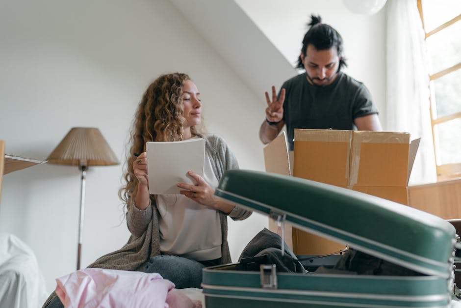 Inside a residential room during home relocation, a woman with curly hair is seated and inspecting a white cardboard box, surrounded by packing materials. An open suitcase with a green exterior and metallic locks is on the bed in front of her, partially filled with clothing or linens. Behind her, a man with dark hair tied back, dressed in a dark t-shirt, stands near a cardboard box on a surface, making a peace sign with his fingers. The room is lit by natural light coming through a window covered with sheer curtains, creating a bright environment. In the background, a wooden bedside lamp with a fabric lampshade is visible, along with some additional packing items. This scene captures the process of packing and preparing belongings for a house move, with clothing, cardboard boxes, and packing equipment indicating an ongoing furniture transport operation, consistent with professional removals by [COMPANY_NAME], as referenced on the page for removals near Chessington World of Adventures.
